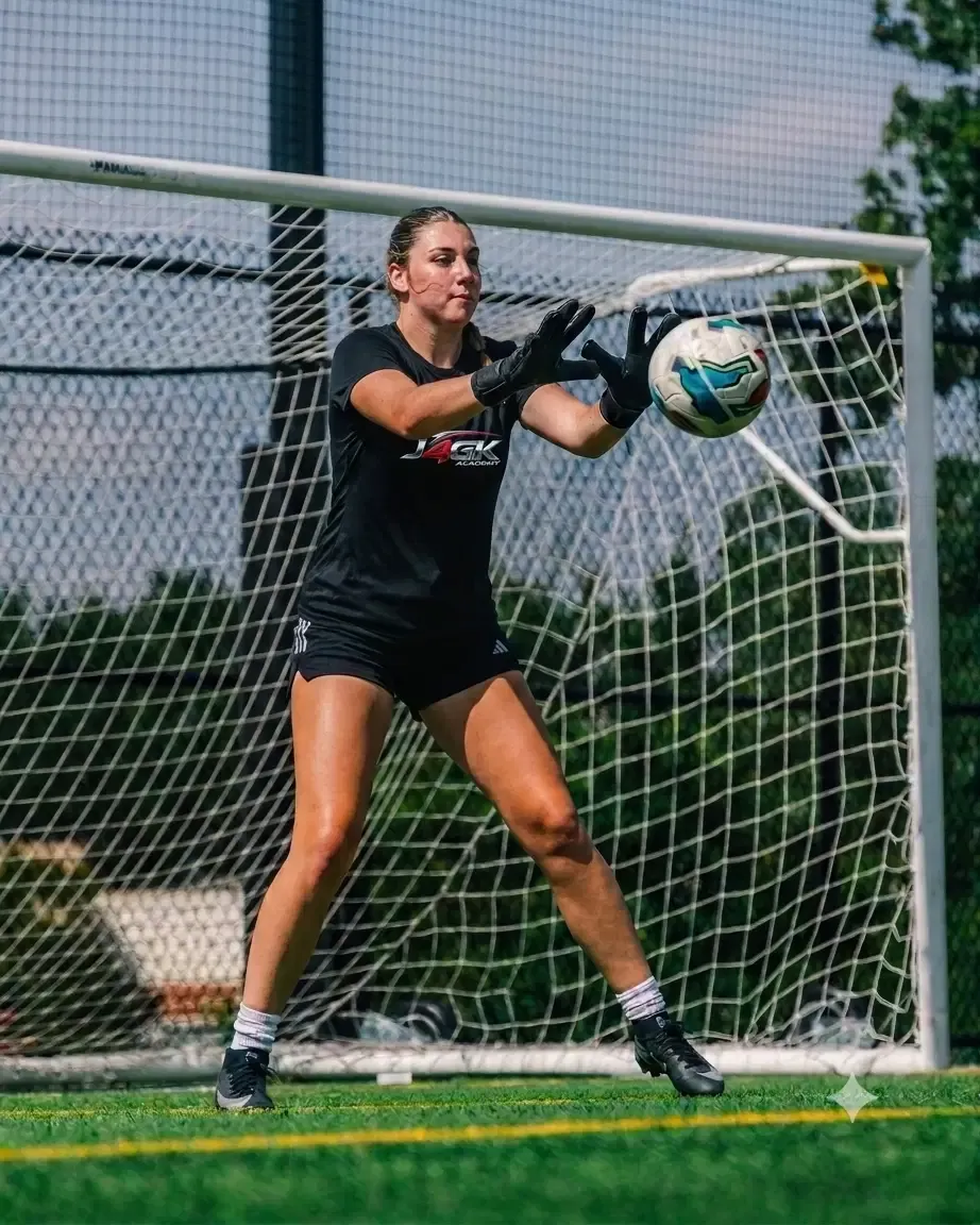 Young female goalkeeper catching a ball in front of a goal in a Just4GK shirt