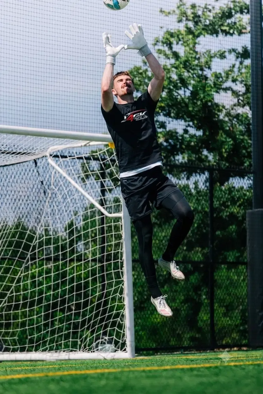 Male goalkeeper jumping to catch a high ball at the goal line in a Just4GK shirt